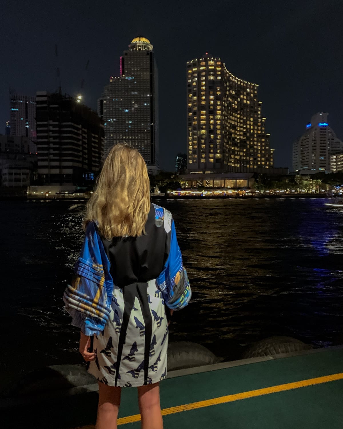 Cec in a bold, patterned dress stands by the river at night, gazing at Bangkok’s lit-up skyline and city reflections.