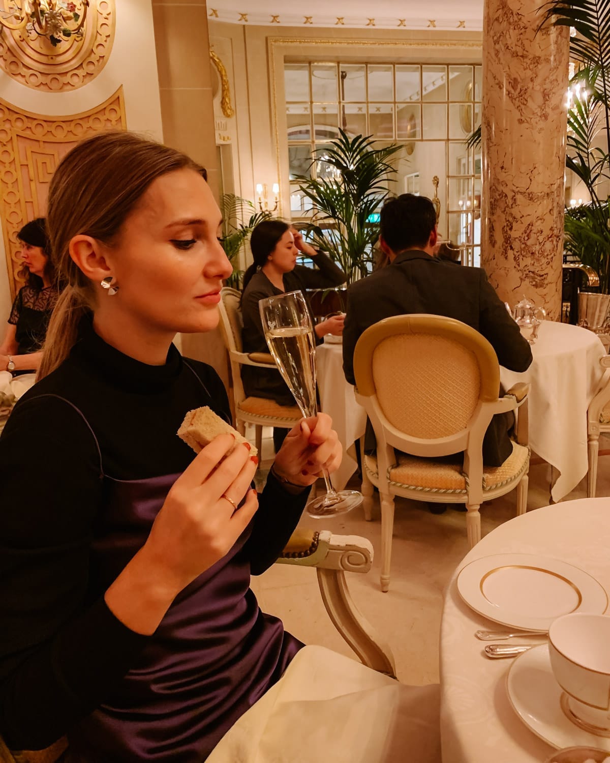 Cec dressed up enjoying afternoon tea in a lavish tea room, holding a champagne flute and tea sandwich in London.