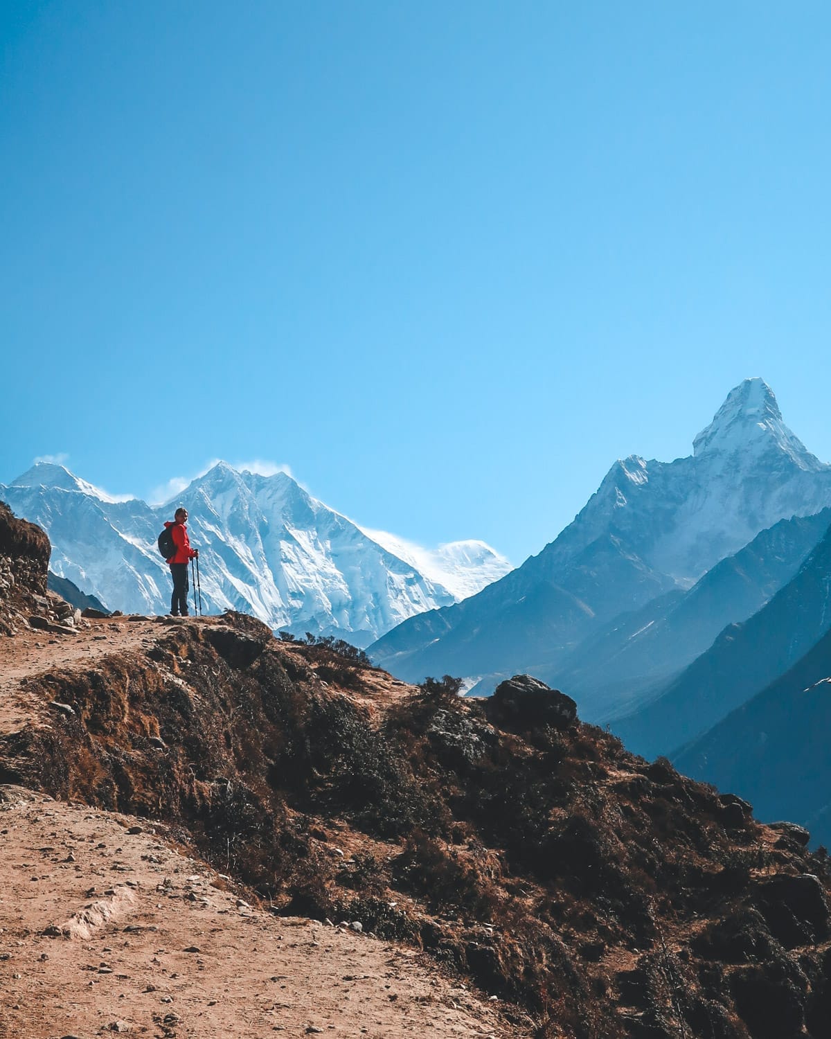 Cec with trekking poles stands on a mountain path, dwarfed by the massive snow-covered peaks of the Himalayas.