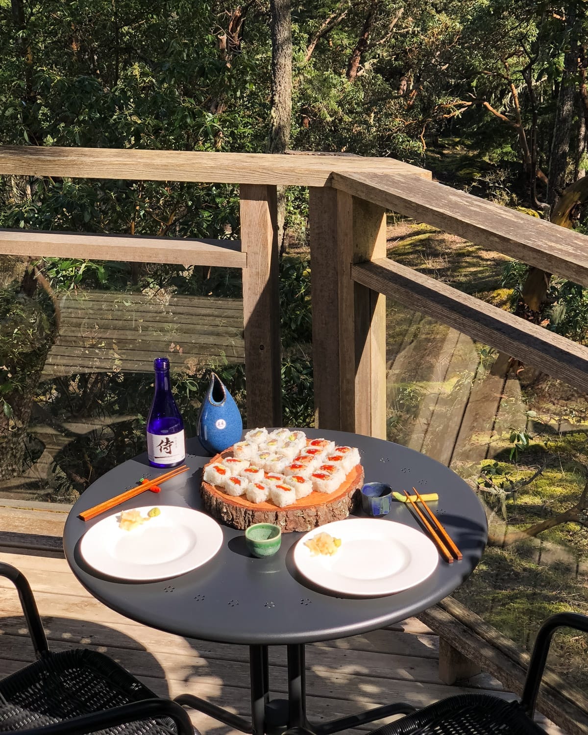 Outdoor table set with sushi rolls from Express Sushi 2 Go, sake, two plates and chopsticks, surrounded by lush forest views on Salt Spring Island.