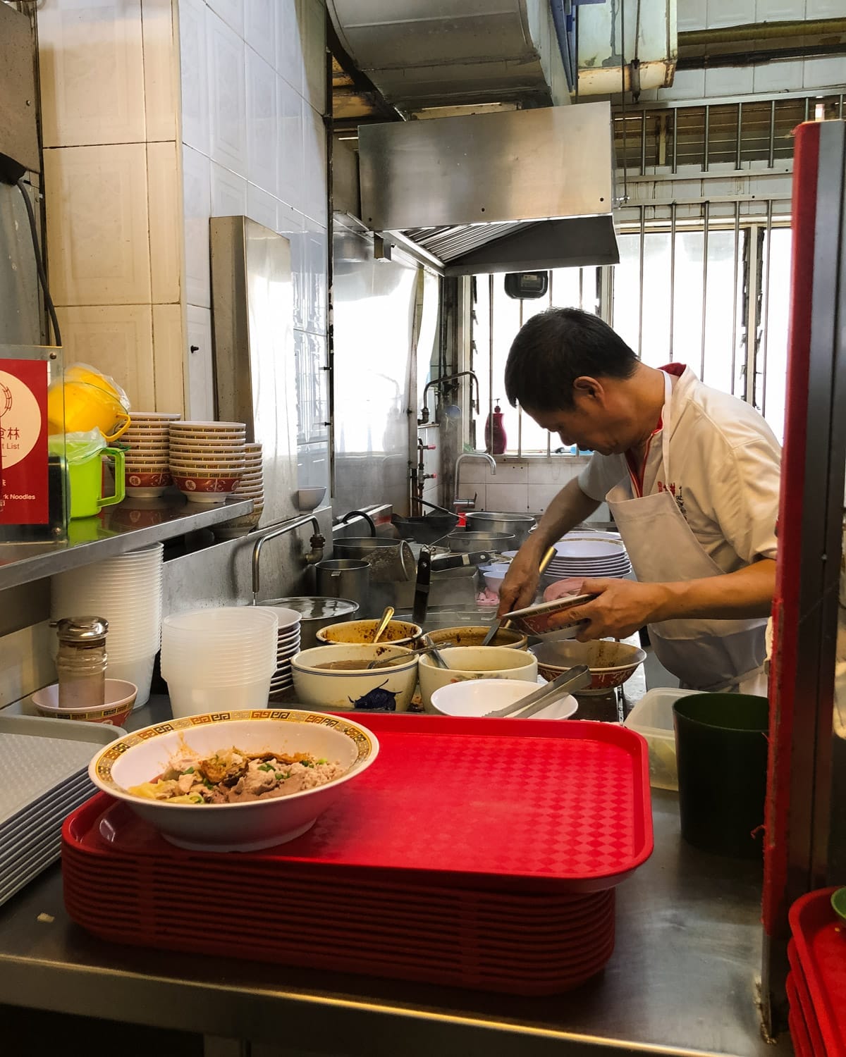 A chef preparing bowls of noodles in a traditional Singapore hawker stall kitchen.