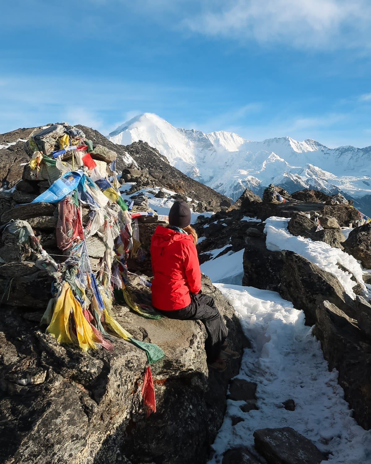 Cec in a red jacket sitting the Gokyo Ri Summit, gazing at snow-covered Himalayan mountains, with prayer flags blowing in the breeze.