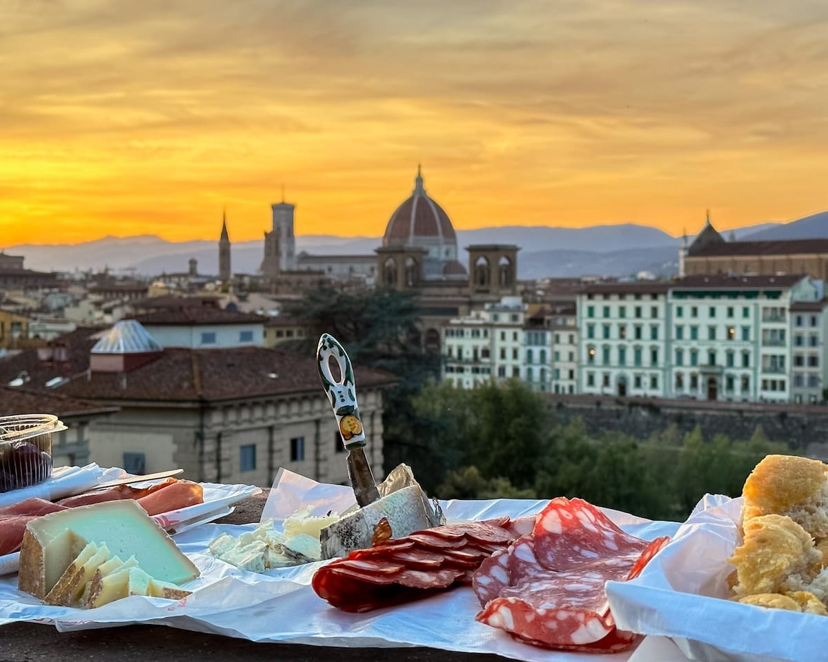 Assorted Italian cheeses and meats laid out picnic-style with Florence’s Duomo and city skyline glowing at sunset.