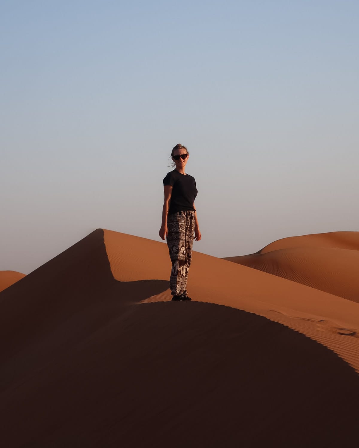 Cec stands on a sunlit sand dune in the Wahiba Sands of Oman, with soft golden ripples stretching into the horizon.