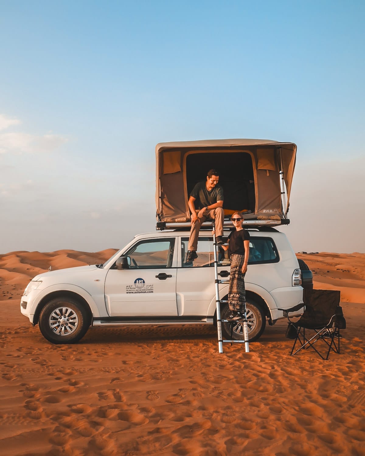 Cec & Ari camping in the desert with a rooftop tent on their SUV, surrounded by endless sand dunes under a golden sky in Oman.