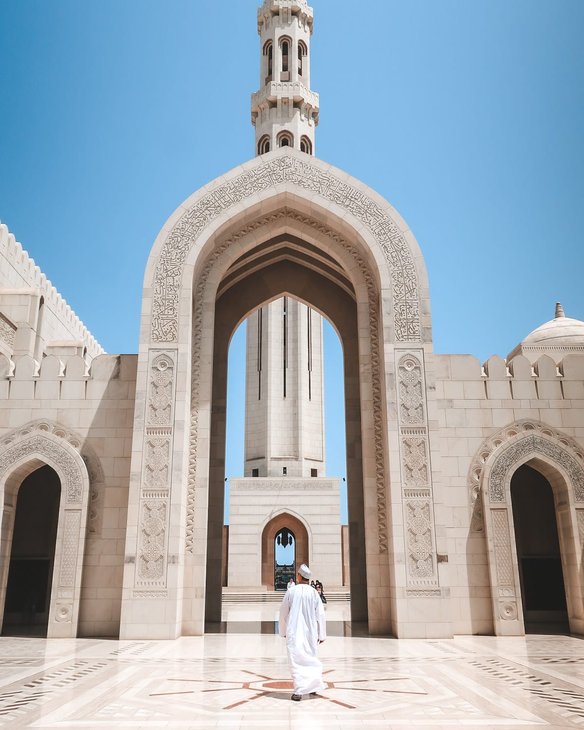 Ari in a white robe walks toward the towering minaret of Sultan Qaboos Grand Mosque, framed by detailed Islamic architecture.