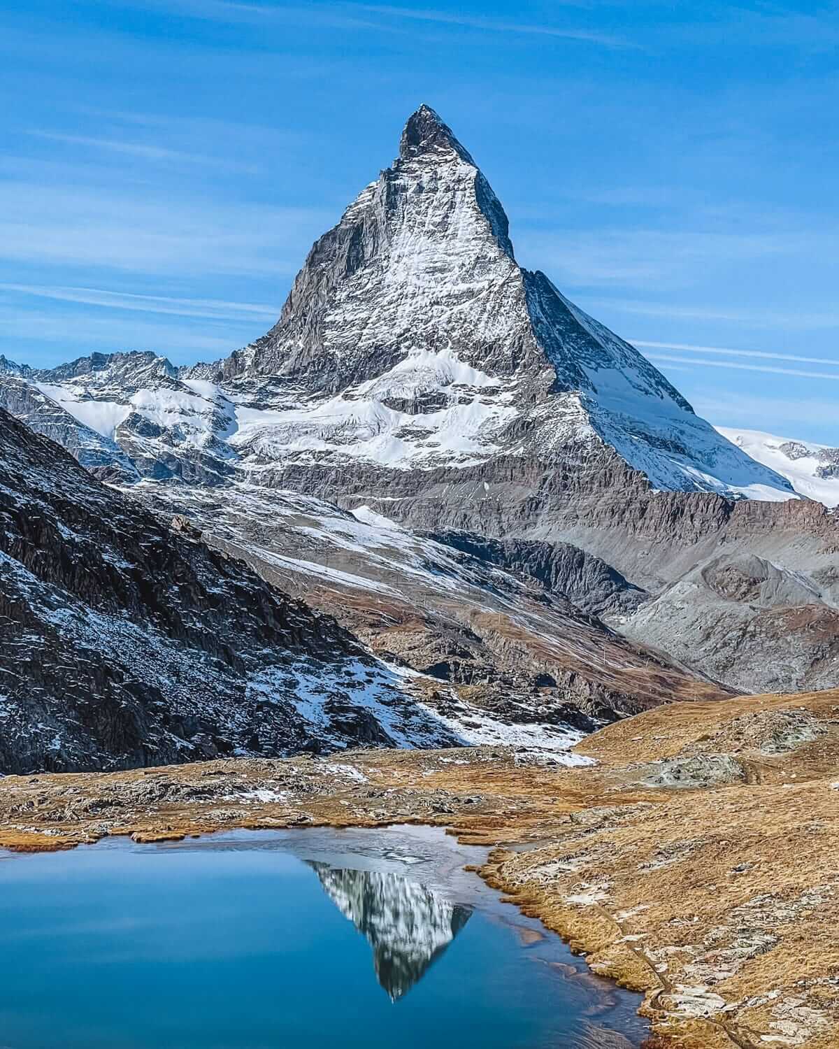 The snow-covered Matterhorn stands tall under a blue sky, perfectly mirrored in a still alpine lake at its base.