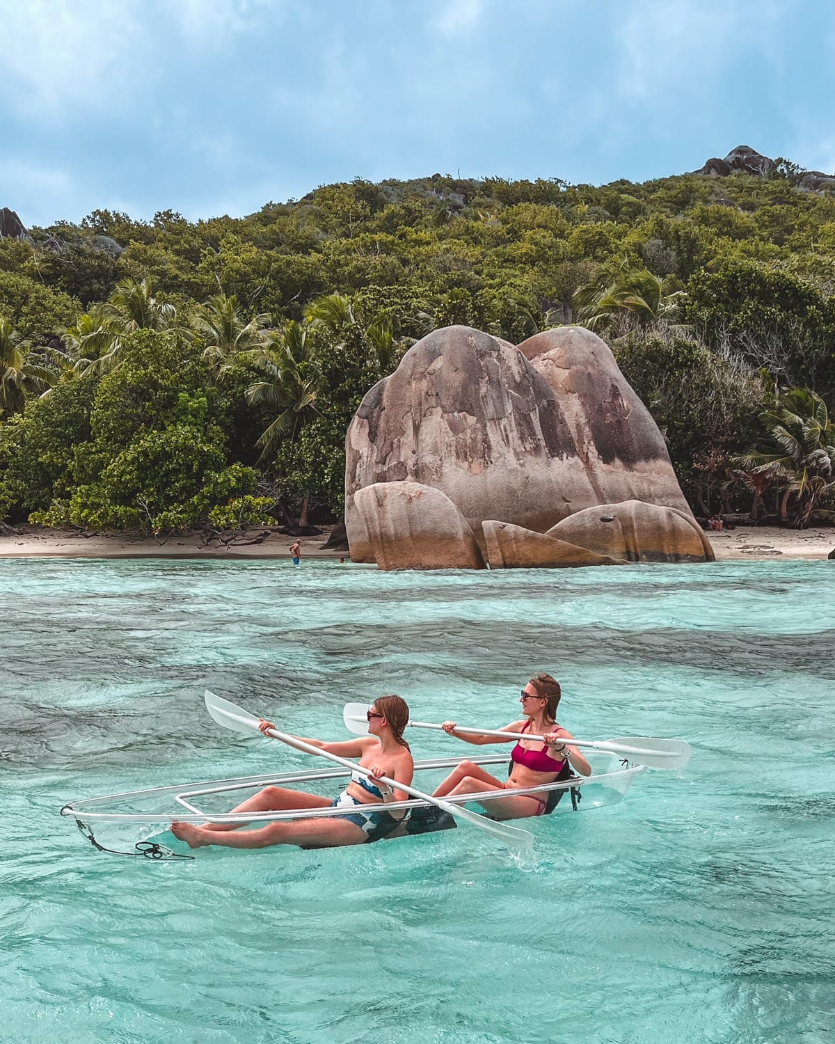 Cec and her sister paddling a clear kayak through turquoise waters along the granite boulders and jungle coast of Seychelles.