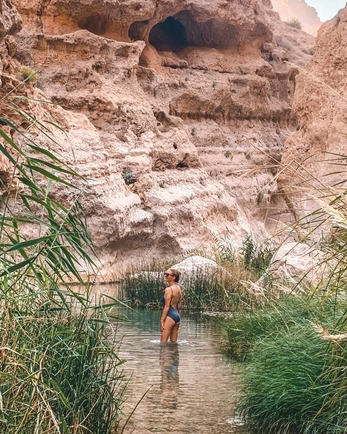 Cec wades through a clear desert wadi surrounded by tall grasses and rugged red rock cliffs in Oman.