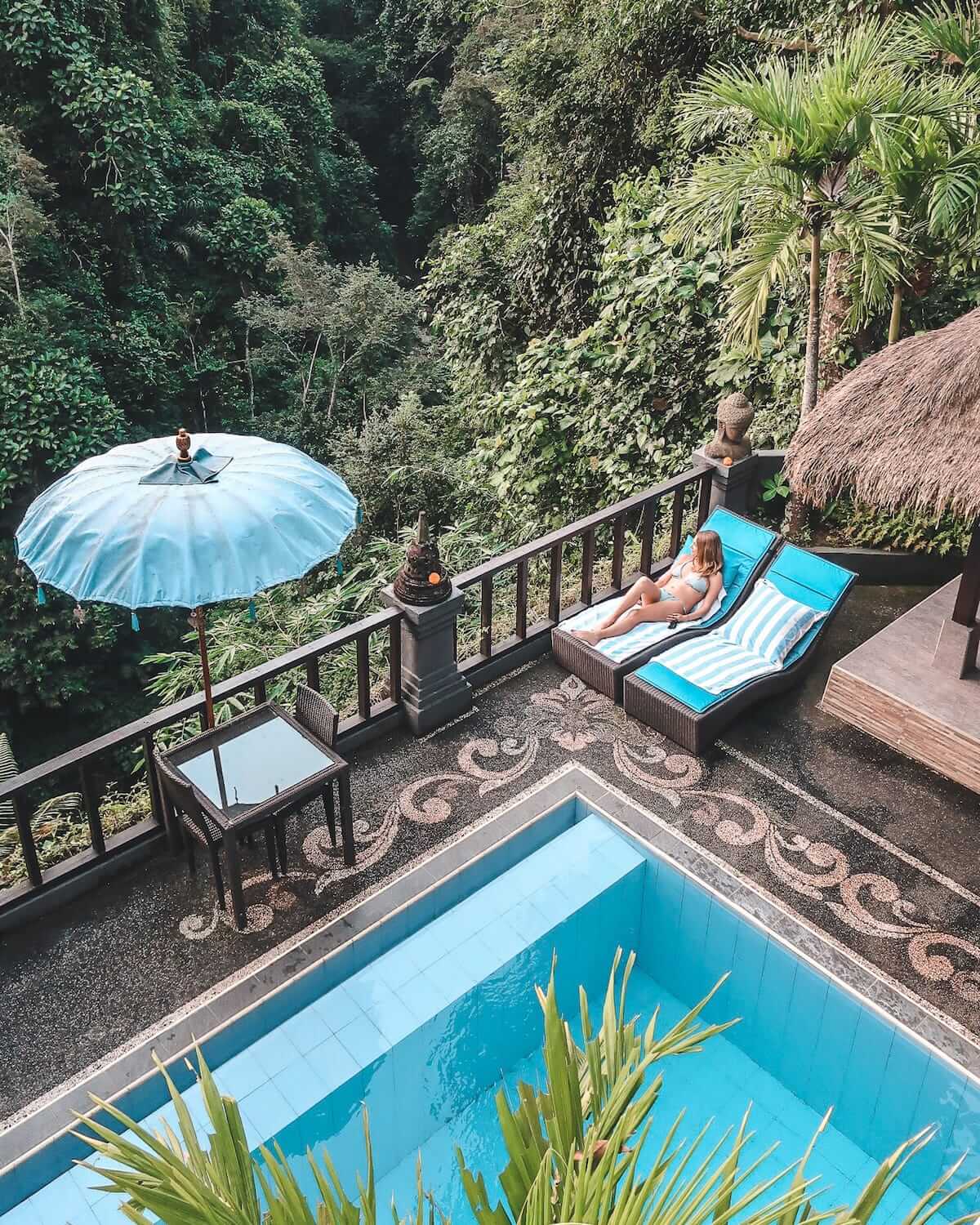 Cec lounging on a blue lounger beside pool in a lush jungle setting, with tropical plants and a blue umbrella nearby.