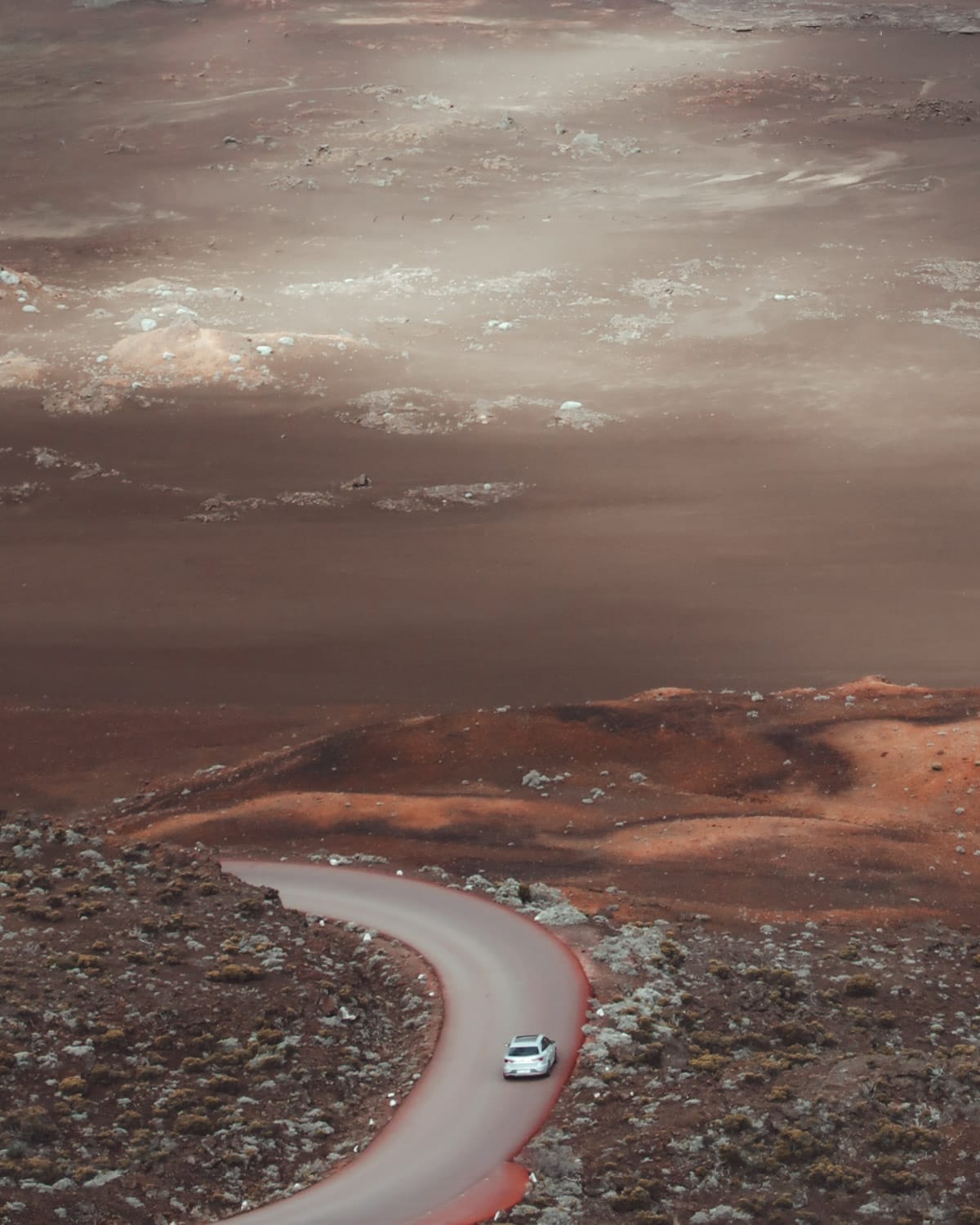 A white car drives a winding road through a reddish-brown volcanic landscape on the Route du Volcan in Réunion.