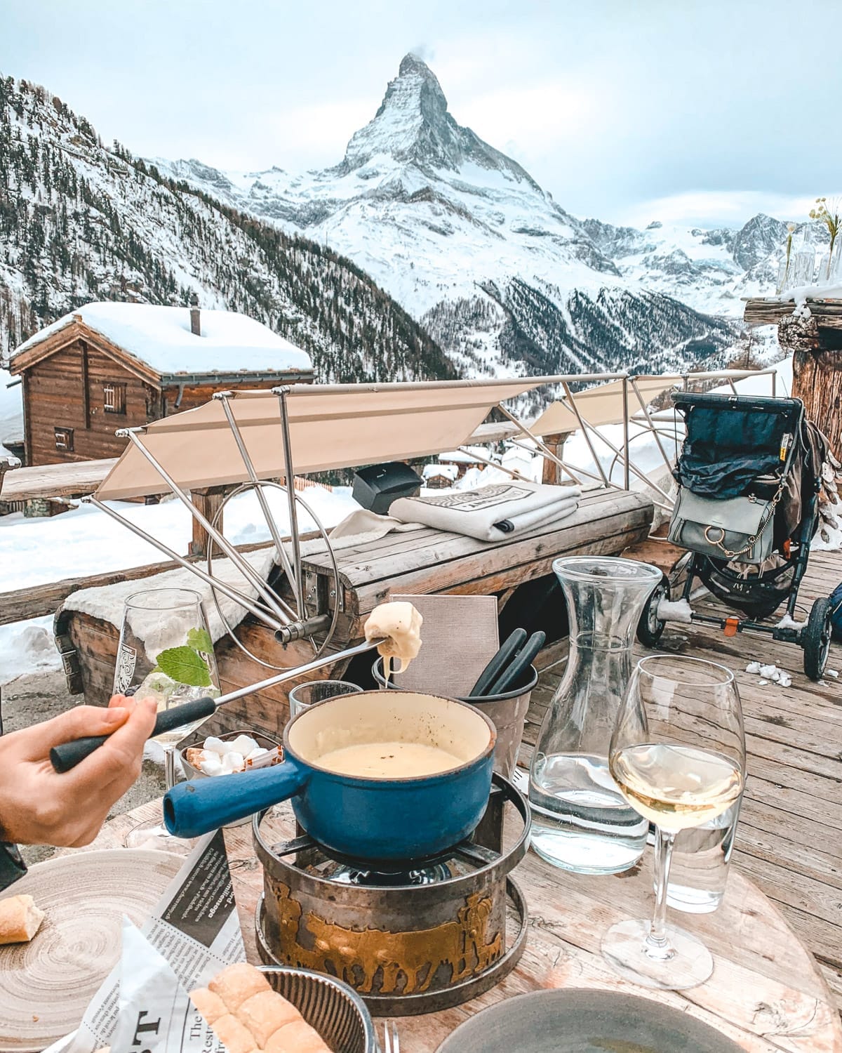 Outdoor cheese fondue meal set against the snowy Matterhorn in Zermatt, with wine glasses and cozy mountain lodge vibes.