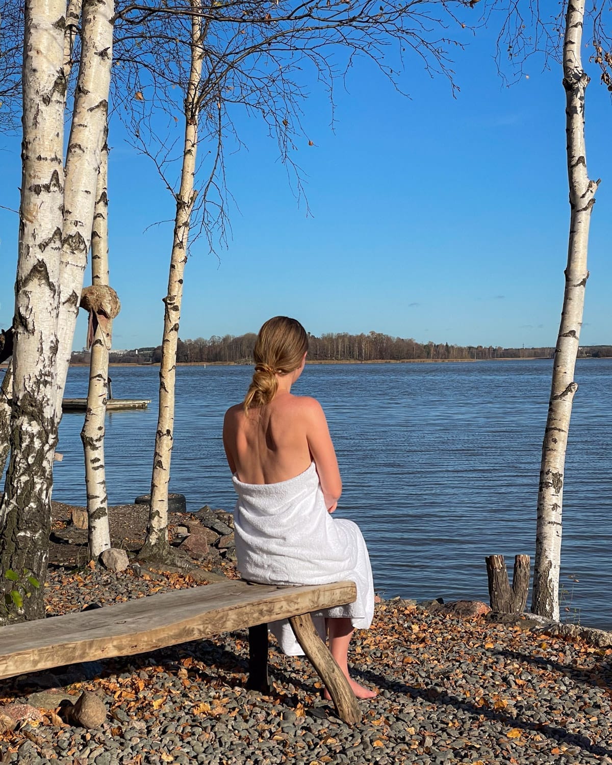 Cec in a towel sits on a bench by a lake after a traditional Finnish sauna, surrounded by birch trees.