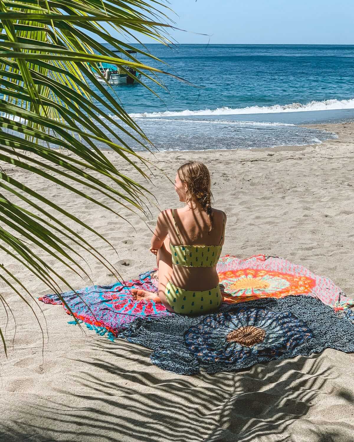Cec in a yellow bikini sits on a colourful beach towel under palm fronds, gazing out at the blue ocean waves in St. Lucia.