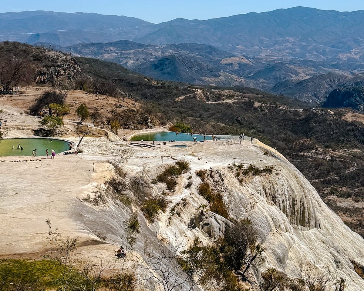 View of Hierve el Agua’s natural rock formations and infinity pools on a cliffside in Oaxaca, with vast mountains in the distance.