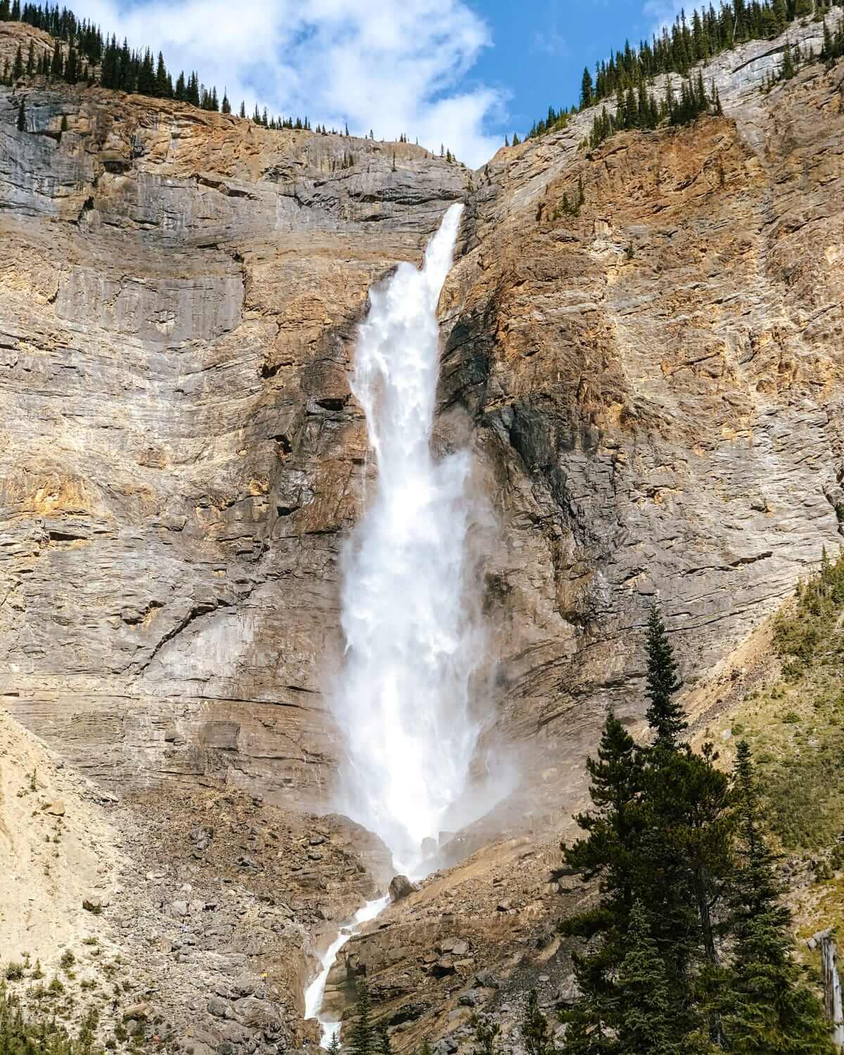 Tall waterfall cascades down a steep rock face at Takakkaw Falls in Yoho National Park, framed by trees and blue sky.