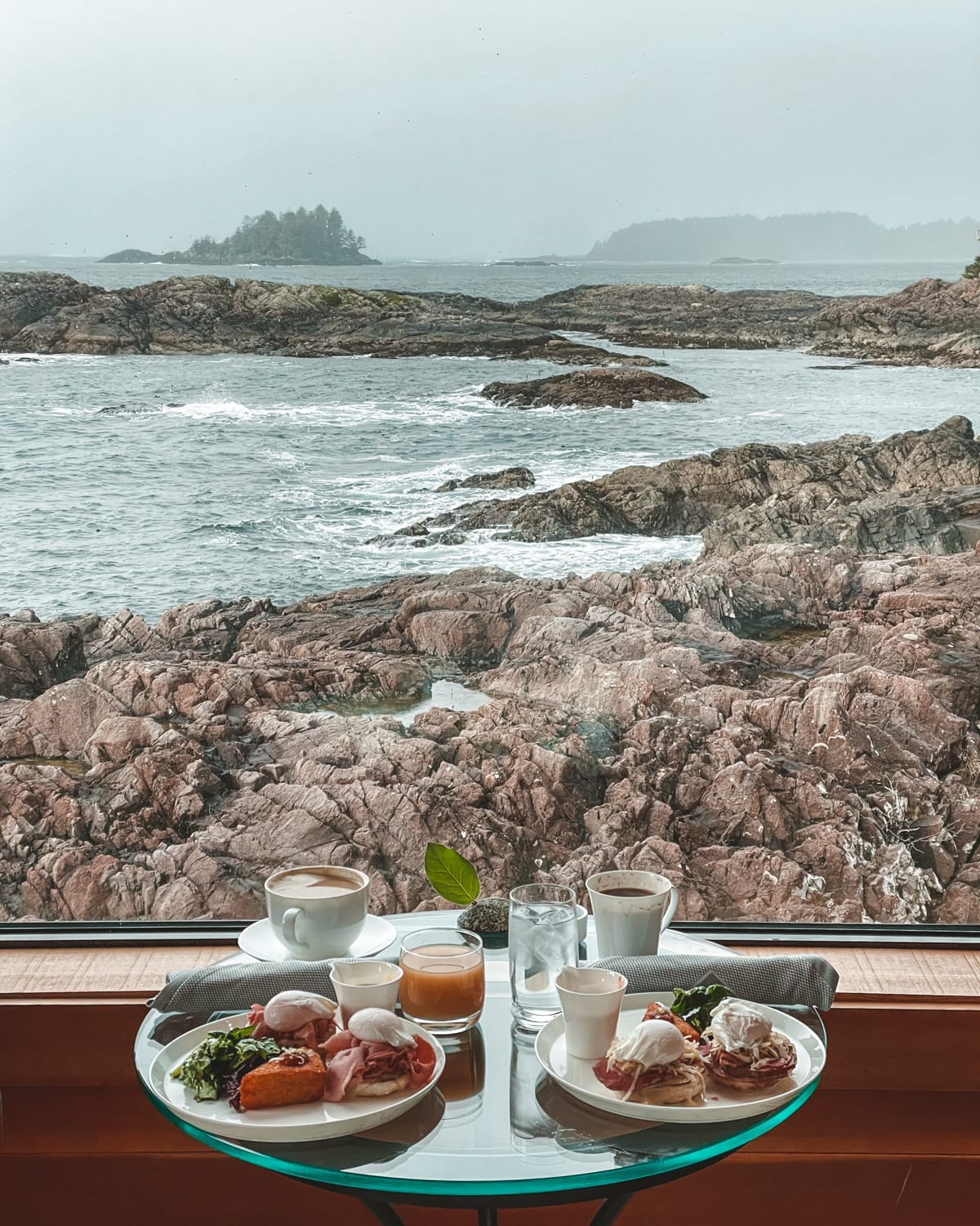 A table set with coffee, juice, and eggs benedict overlooks a rocky coastline and distant forested islets on a stormy day in Tofino.