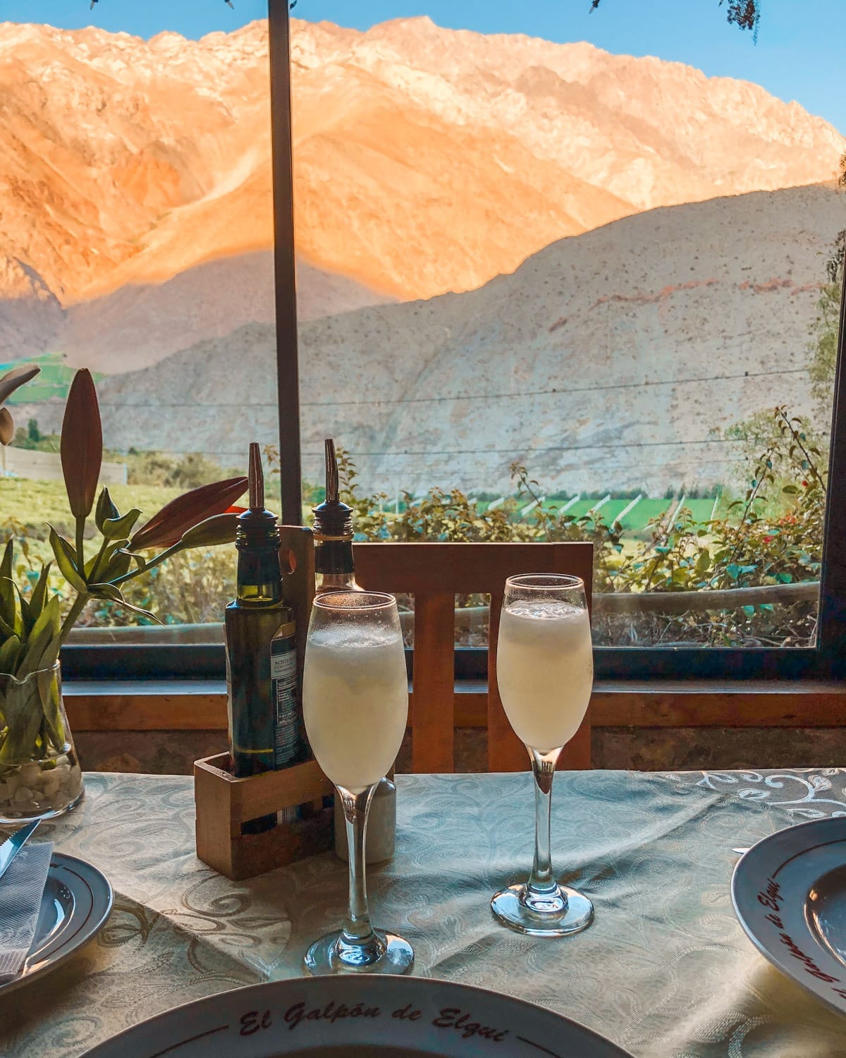 Two glasses of pisco sours on a table at sunset with views of Pisco Elqui’s golden mountains through a window.