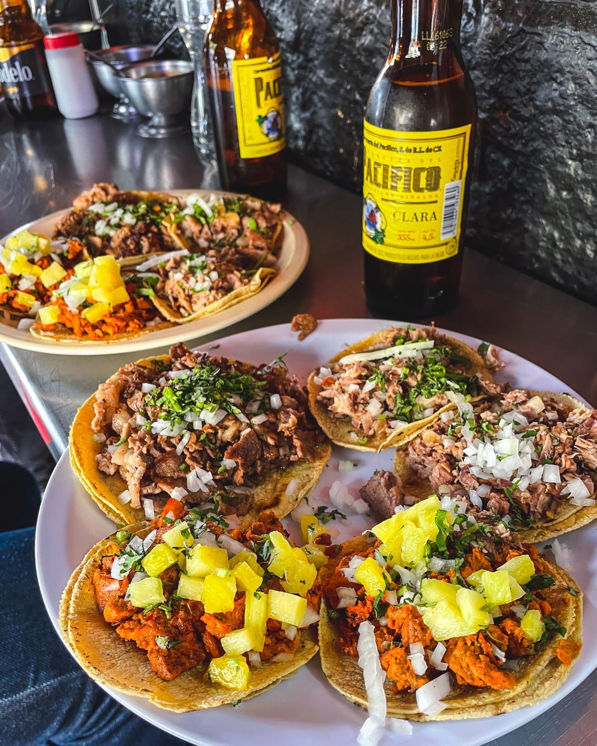 Plate of tacos al pastor and carnitas topped with pineapple, onion, and cilantro, served with cold bottles of Pacifico beer.