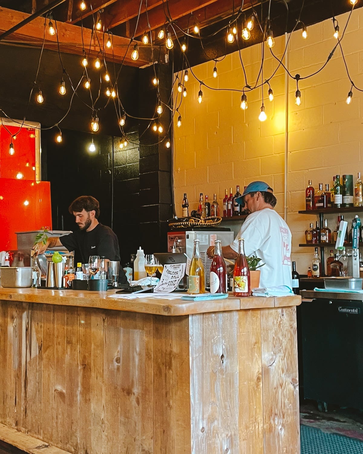 Bartenders preparing drinks at Cassette’s rustic wooden bar, under warm string lights and shelves of spirits.