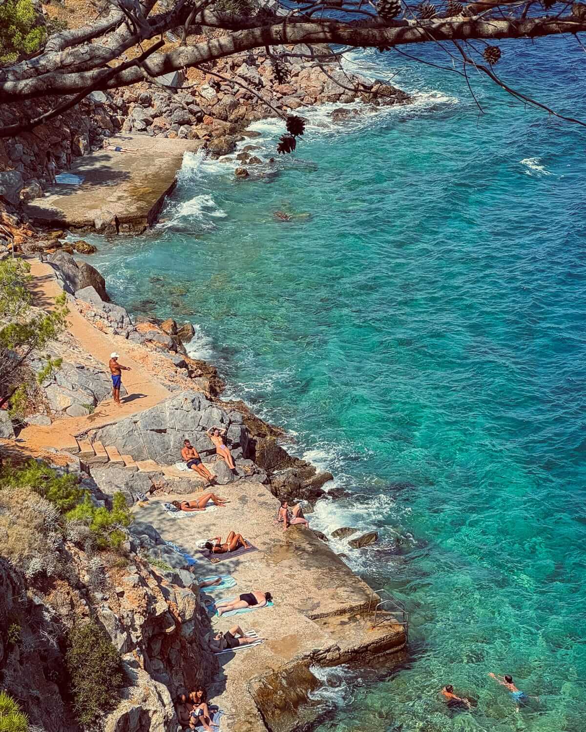 Beach-goers sunbathing and swimming along rocky steps by the turquoise sea in Hydra Island.