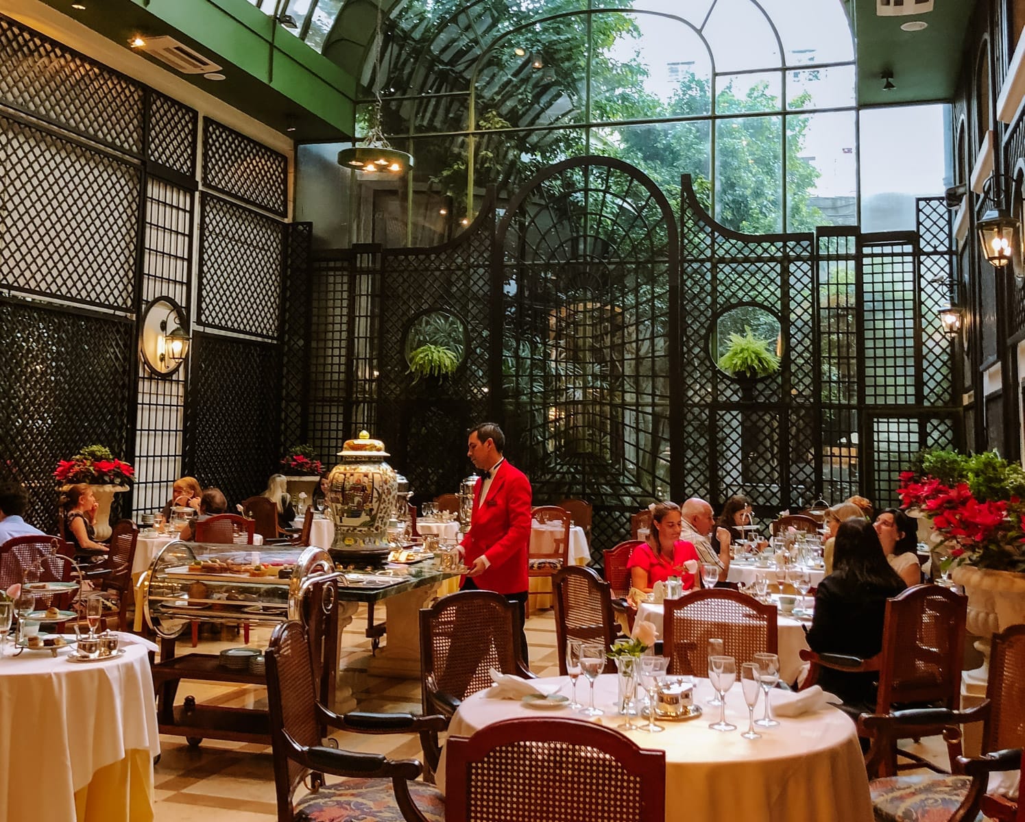 Fine dining room at L'Orangerie with vintage decor, white tablecloths, and a server in a red jacket preparing afternoon tea.