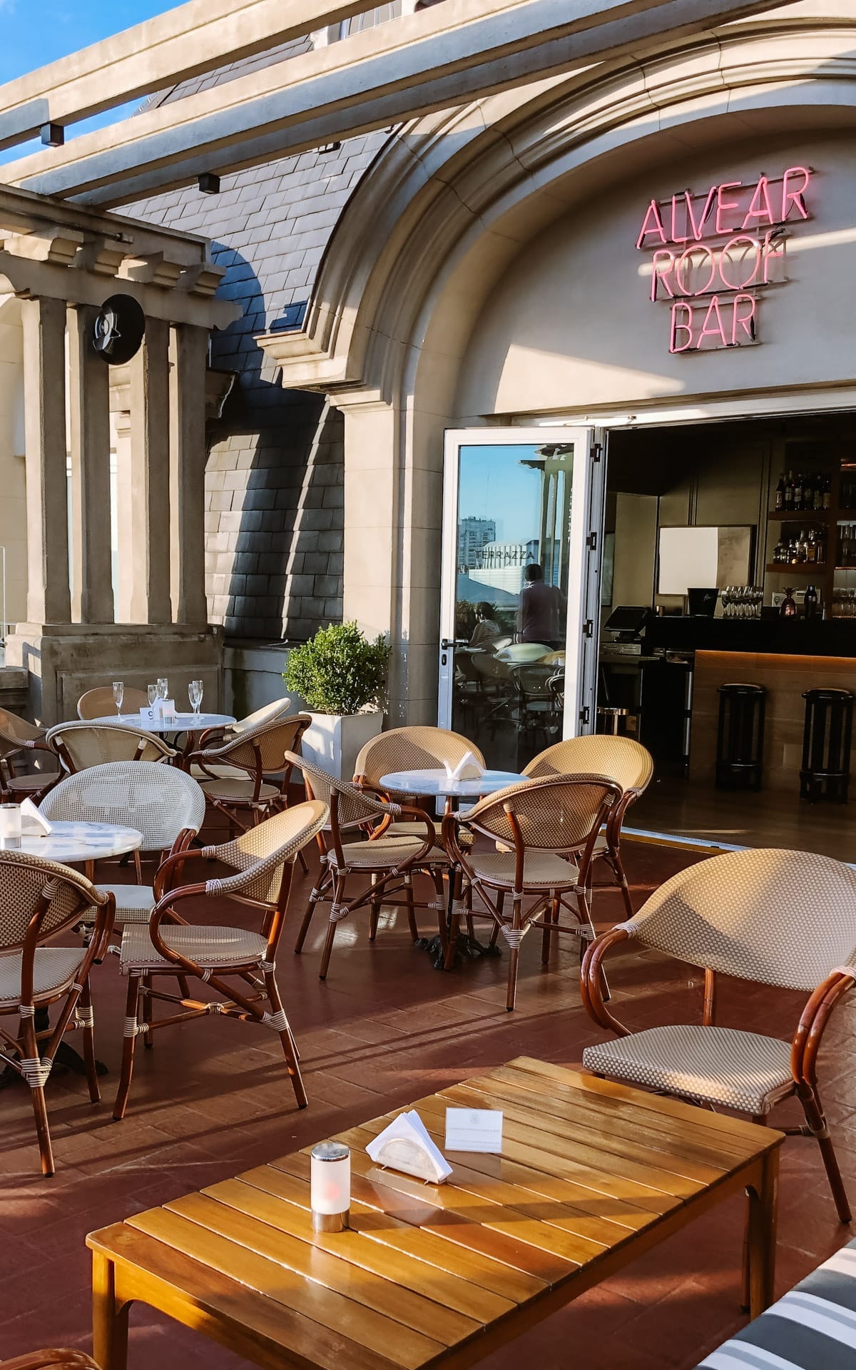 Sunlit rooftop terrace with Parisian-style chairs and a neon Alvear Roof Bar sign above the arched entrance.