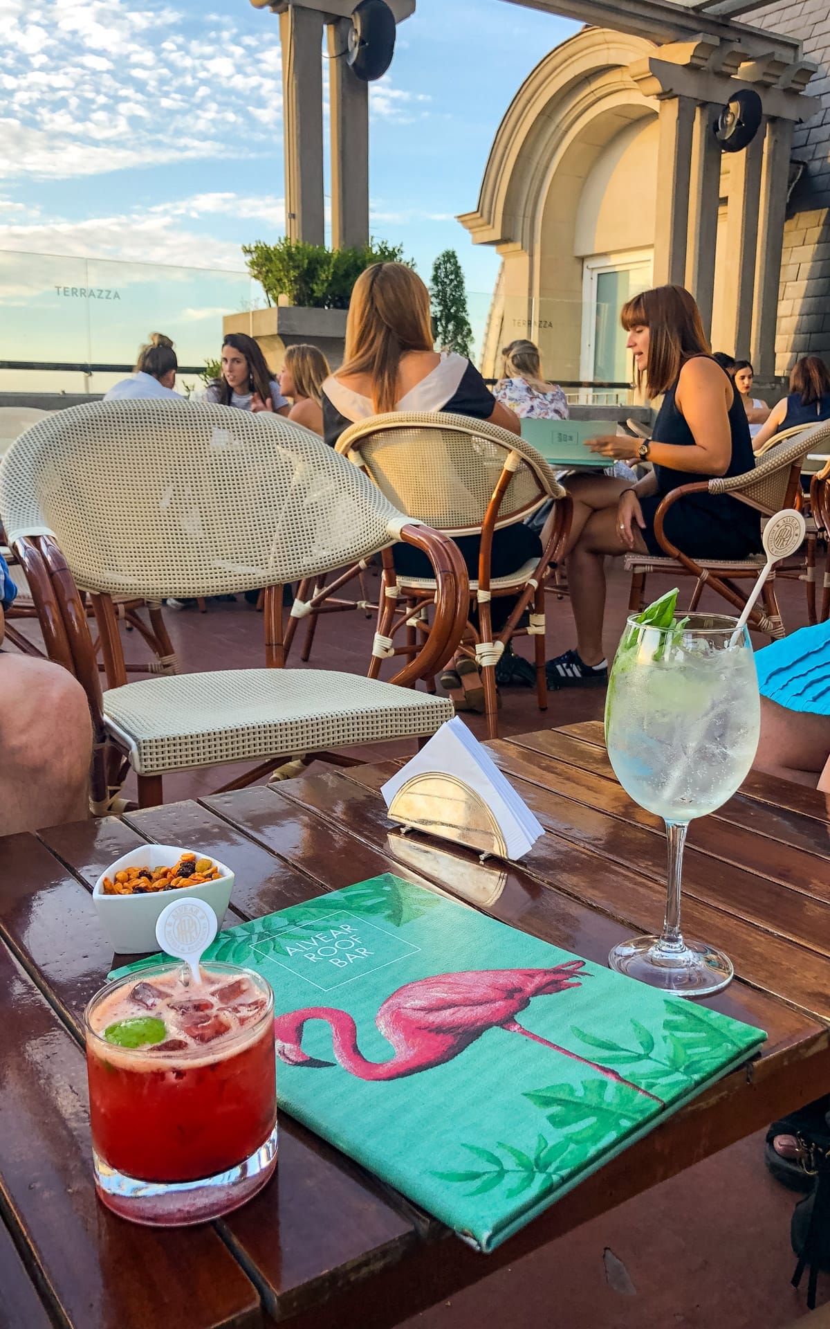 Chic rooftop crowd at golden hour with cocktails on the table and a flamingo-covered drinks menu at Alvear Roof Bar.