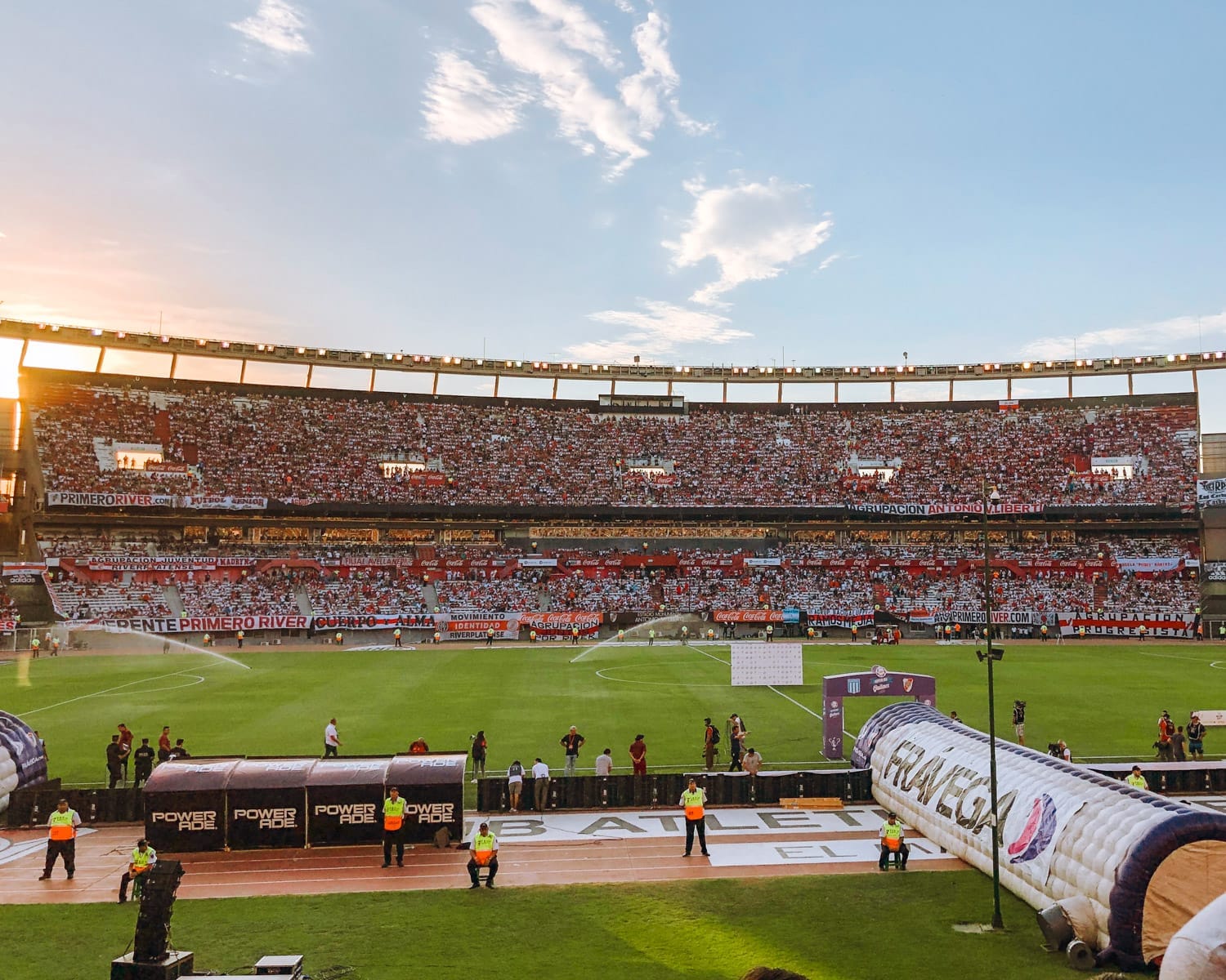 Massive crowd fills the River Plate stadium under a blue sky before kickoff, banners waving and sprinklers running.