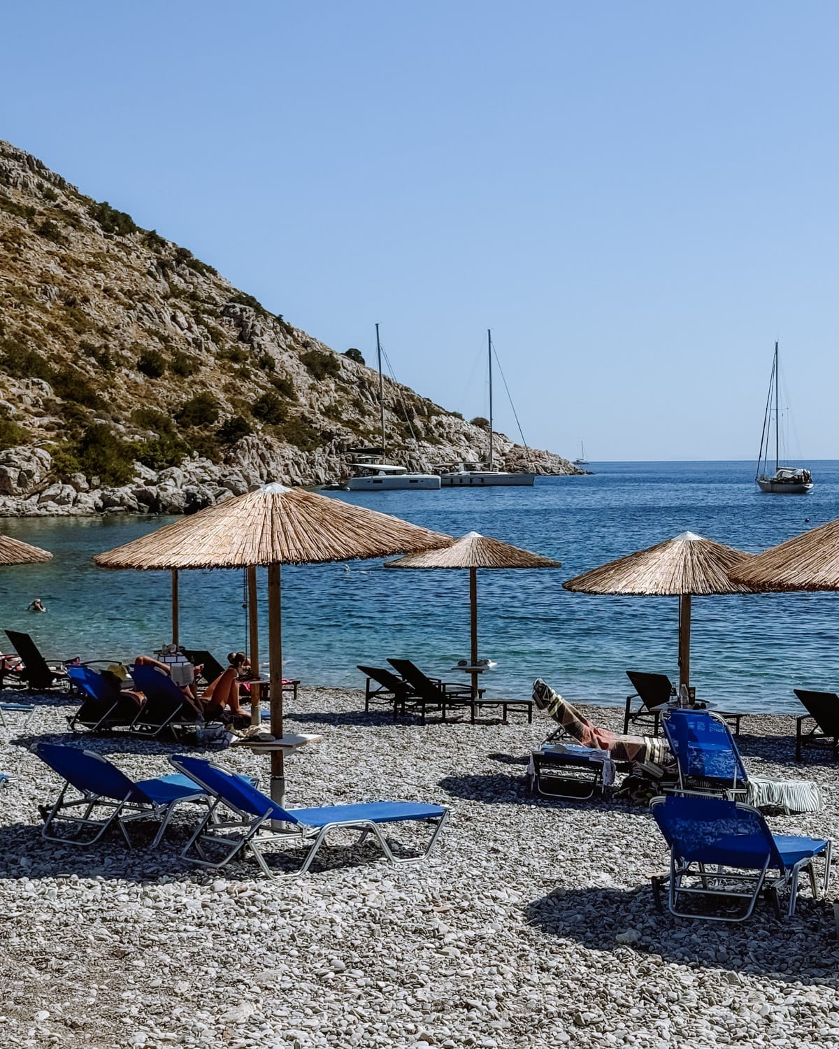 A rocky beach with straw umbrellas and blue sun loungers overlooks a calm bay on Hydra Island, where sailboats float near a rocky hillside.
