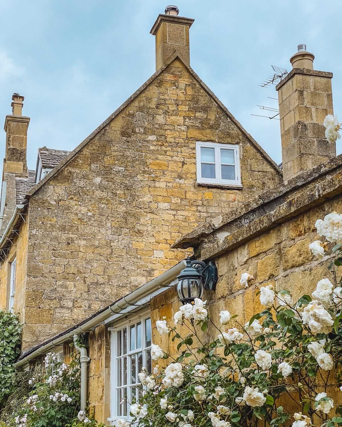 Stone cottage in the English countryside with tall chimneys, white roses, and classic honey-coloured walls.