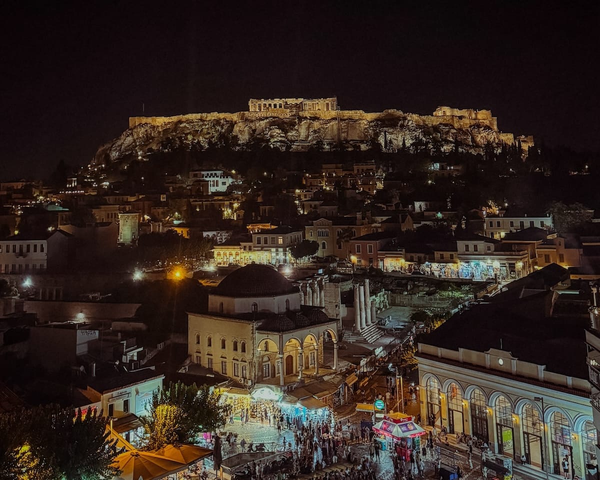 The illuminated Acropolis rises above the lively Plaka district at night in Athens with bustling streets below.