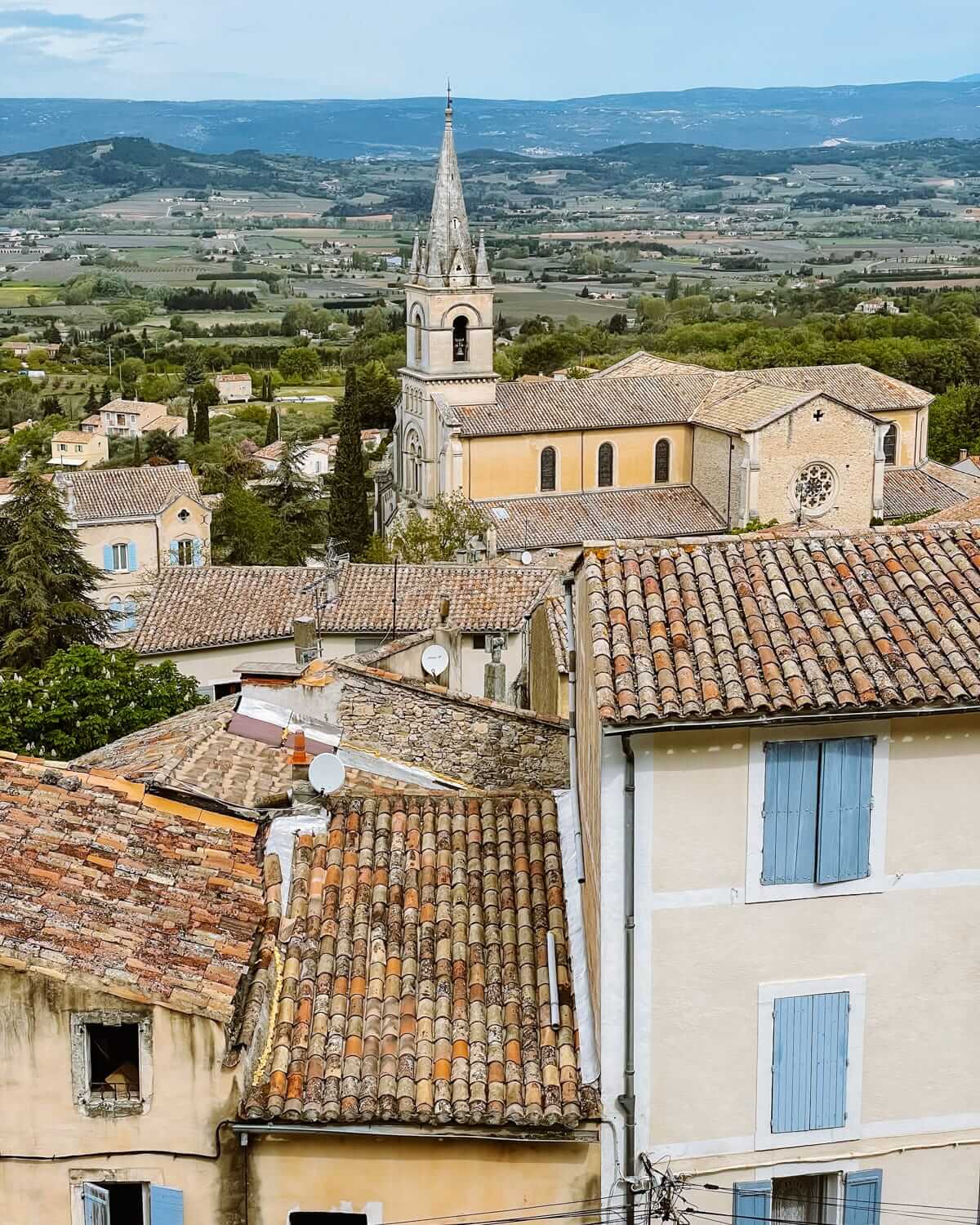 View of church with a pointed steeple among terracotta rooftops in a village overlooking green fields and distant hills in Provence.