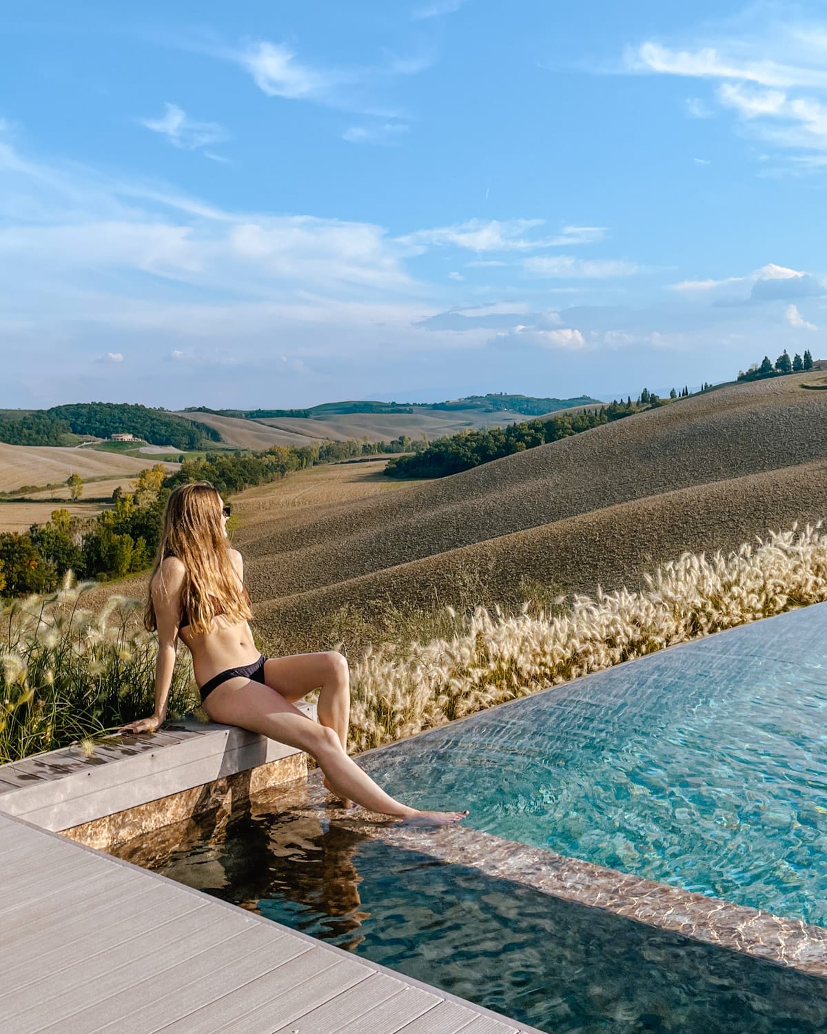 Cec in a bikini relaxes at a infinity pool edge with scenic Tuscan hills, golden grass, and blue sky in the background.