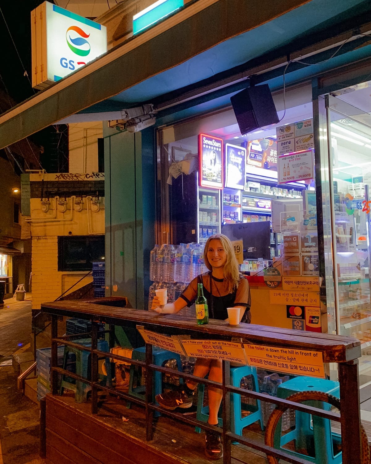 Cec drinking soju outside a Seoul convenience store at night with neon lights in the background.