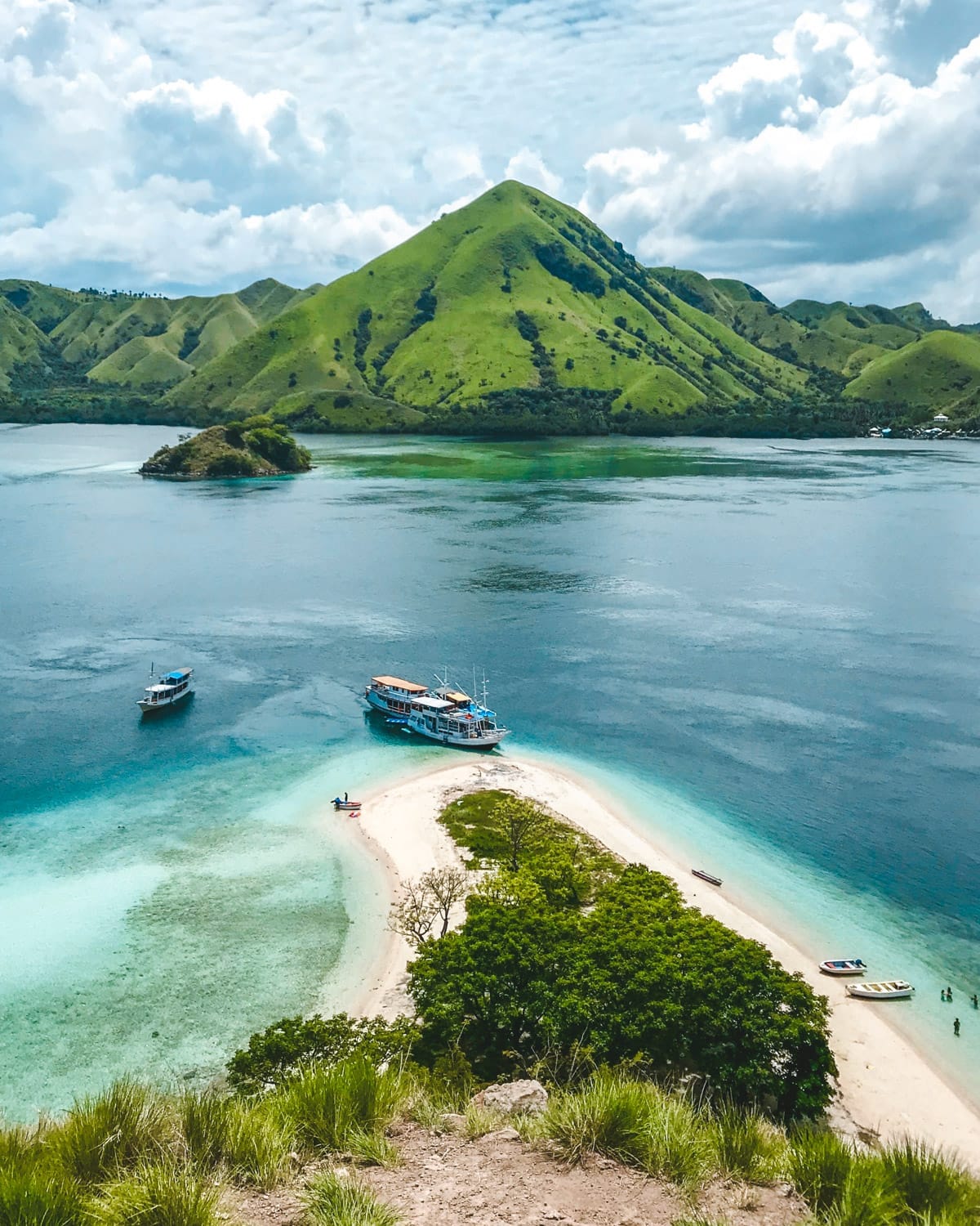 Boats anchored near a white sand spit in Komodo National Park, framed by emerald hills and vibrant blue water.