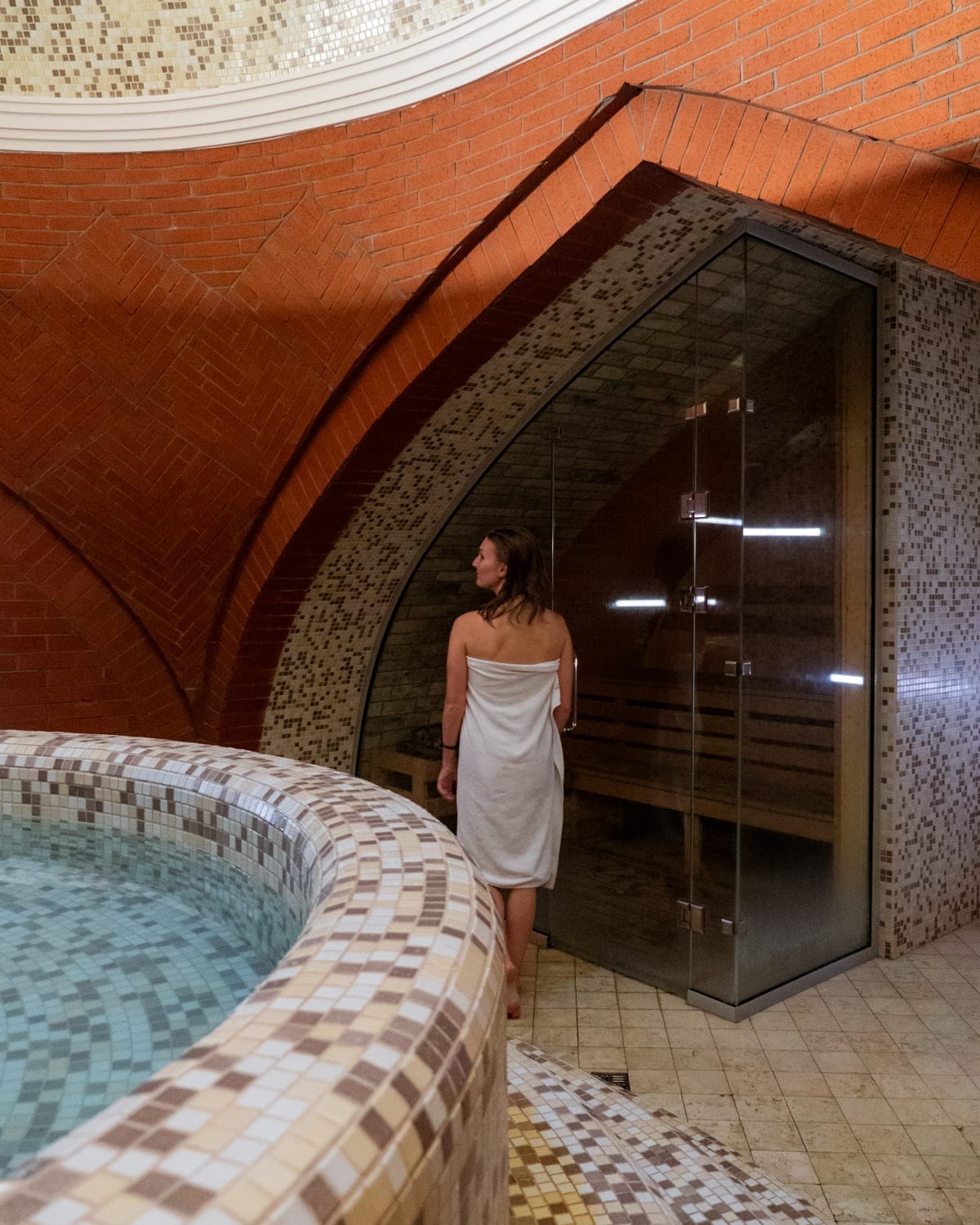 Cec in towel walks into sauna in a tiled thermal spa room in Tbilisi.