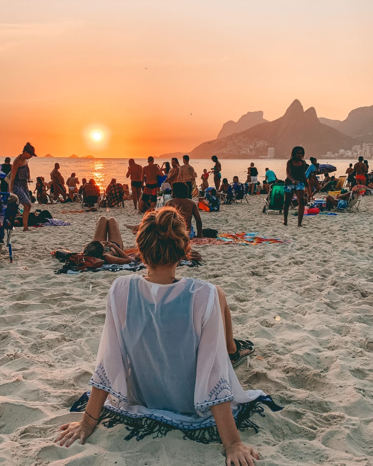 Cec relaxing on Ipanema Beach as the sun sets over the ocean, with a crowd enjoying golden hour and mountains in the background.
