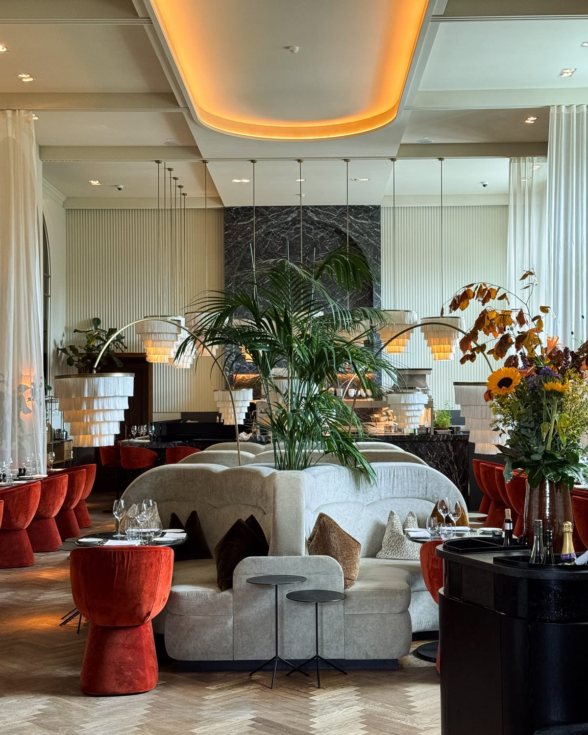 The upscale Madonna restaurant with burnt orange velvet chairs, grey velvet couches, modern chandeliers, large plants, and soft lighting for a chic atmosphere.