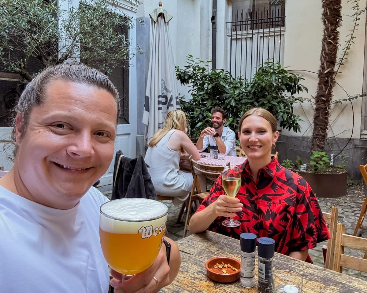 Cec and Ari toasting with beer and wine in the courtyard at Raapstraat 20, surrounded by greenery and other patrons.