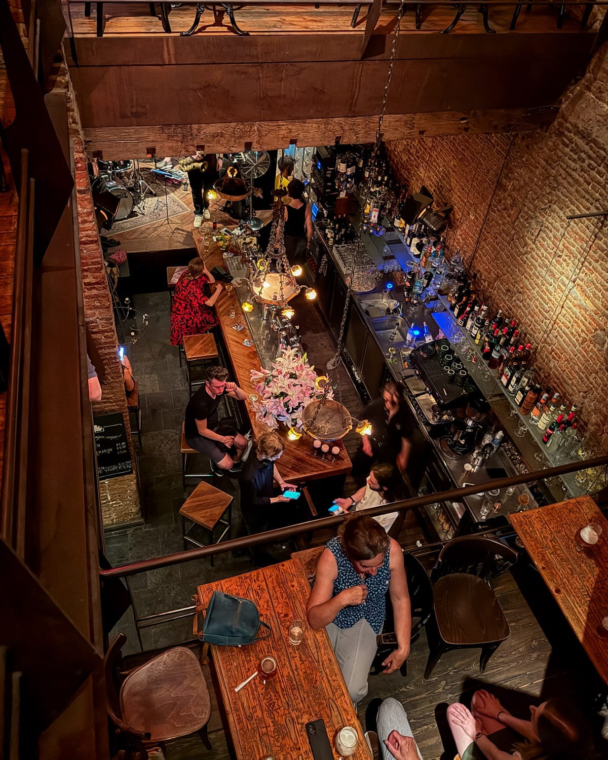 Overhead view of the warm, intimate De Muze Jazz Cafe with exposed brick walls, hanging lights, and people gathered at wooden tables listening to live jazz.