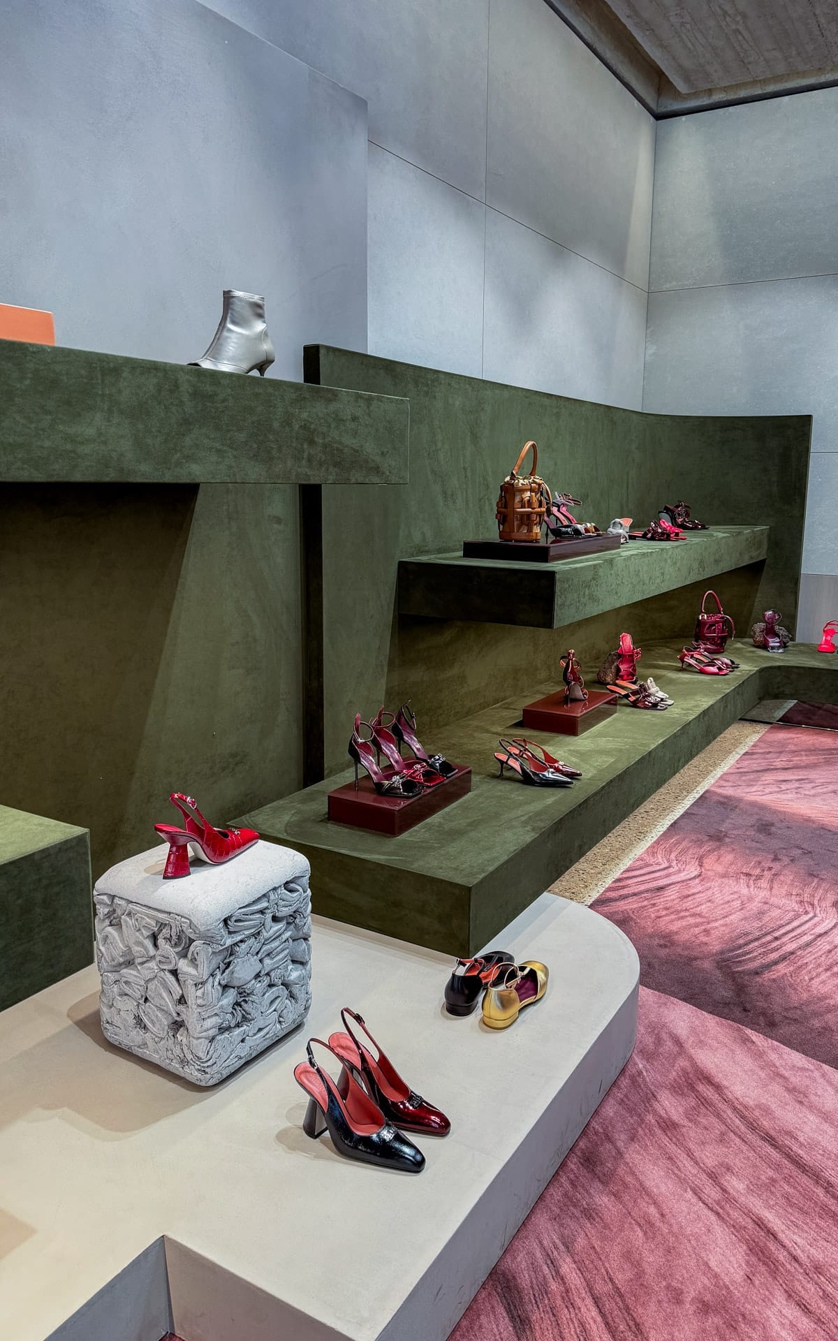 Display of designer shoes and handbags from Morobe on moss-green platforms in a sleek, modern boutique with pink carpet accents.