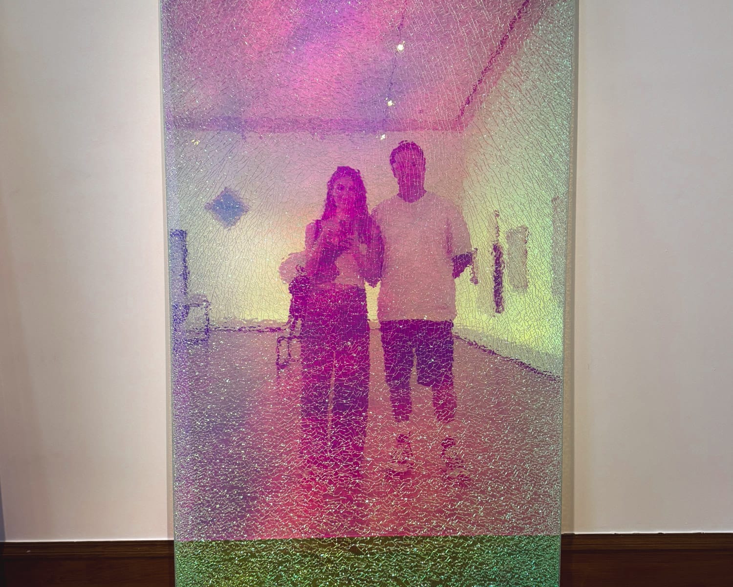 Cec and Ari reflected in a tall, iridescent cracked mirror that glows in pink, purple, and green hues inside KMSKA gallery.