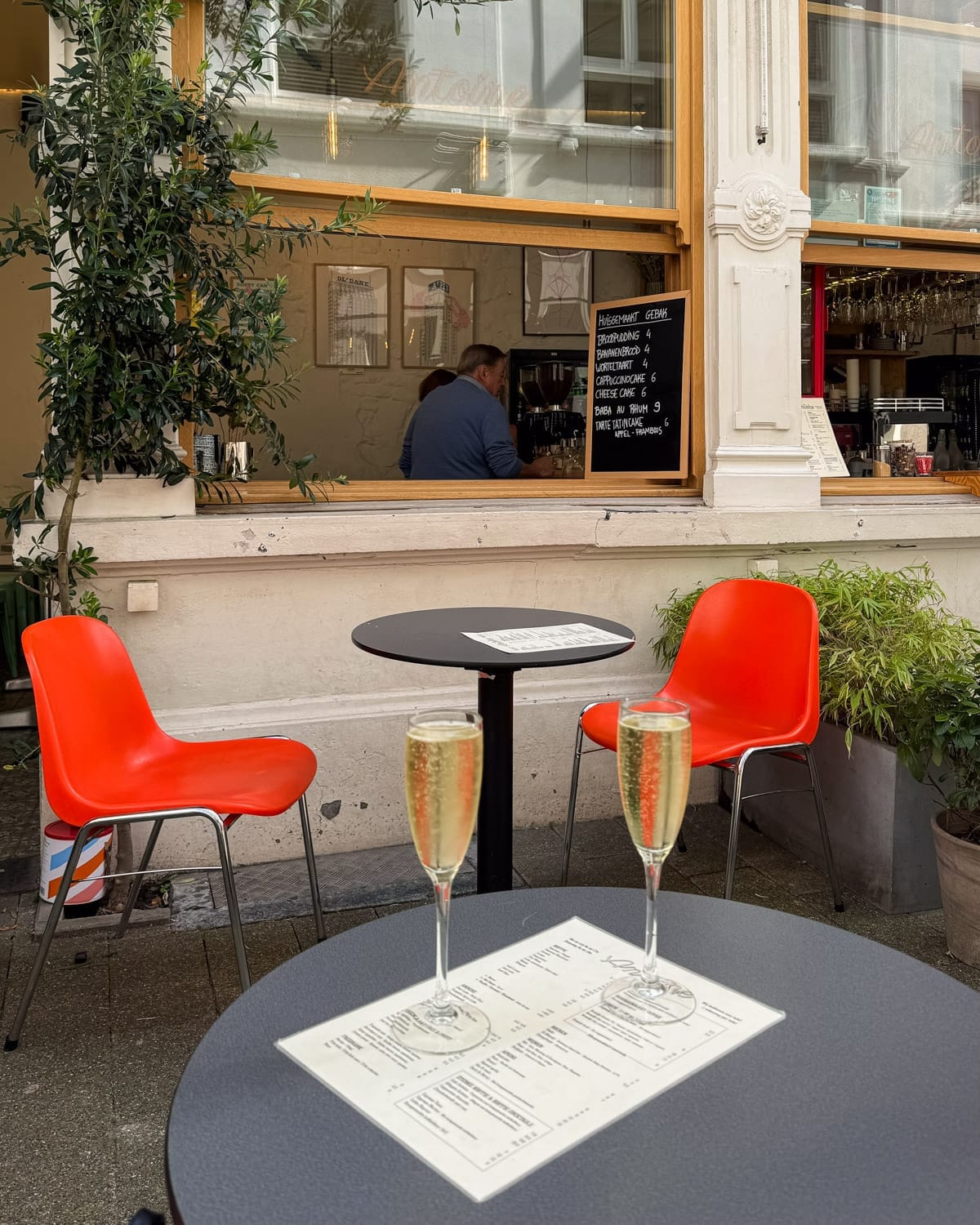 Two glasses of bubbly rest on a black cafe table in front of a cute sidewalk cafe with Aperol orange chairs and leafy plants.