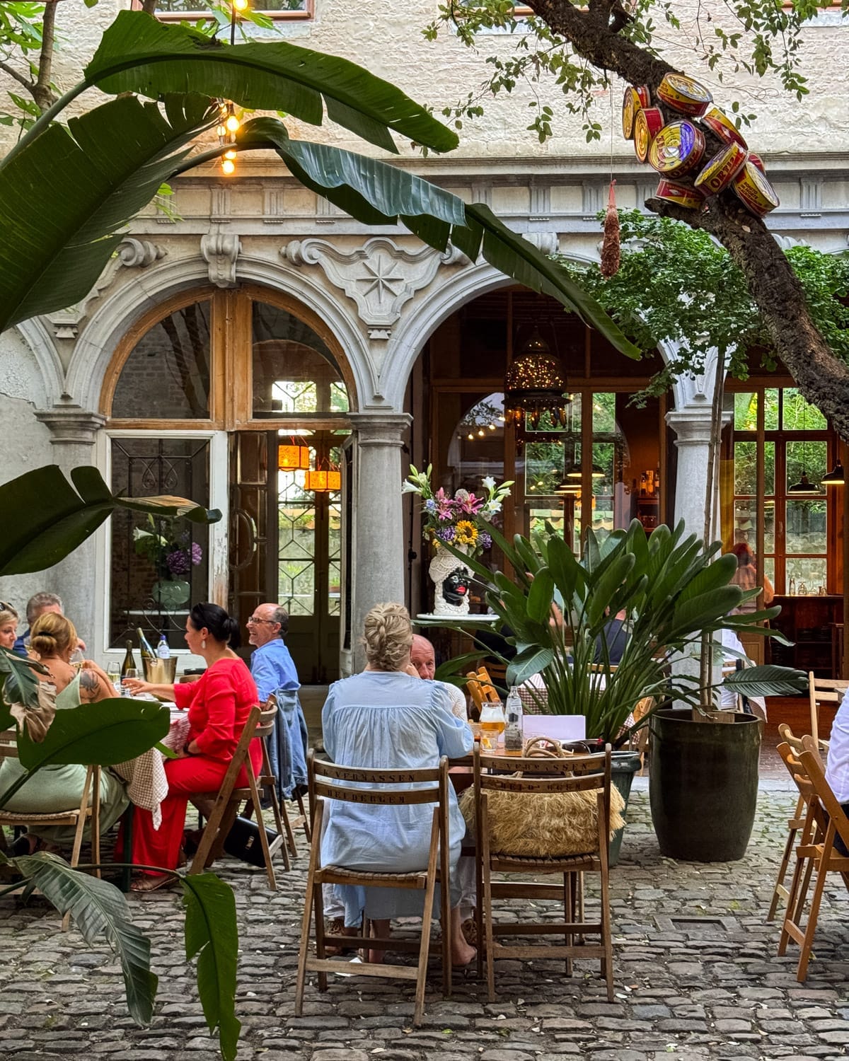 Diners enjoying food and drinks in the cobblestone courtyard of Raapstraat 20 with lush plants and historic arched windows in the background.