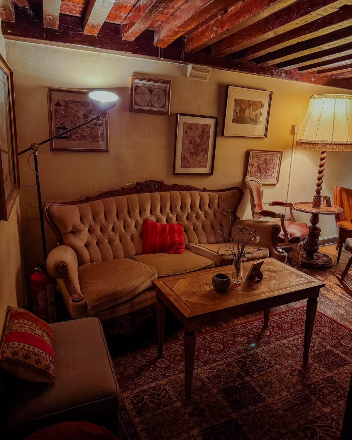 A dimly lit lounge at Jones & Co. with a vintage tufted sofa, eclectic framed art, wooden ceiling beams, and a vintage floor lamp.