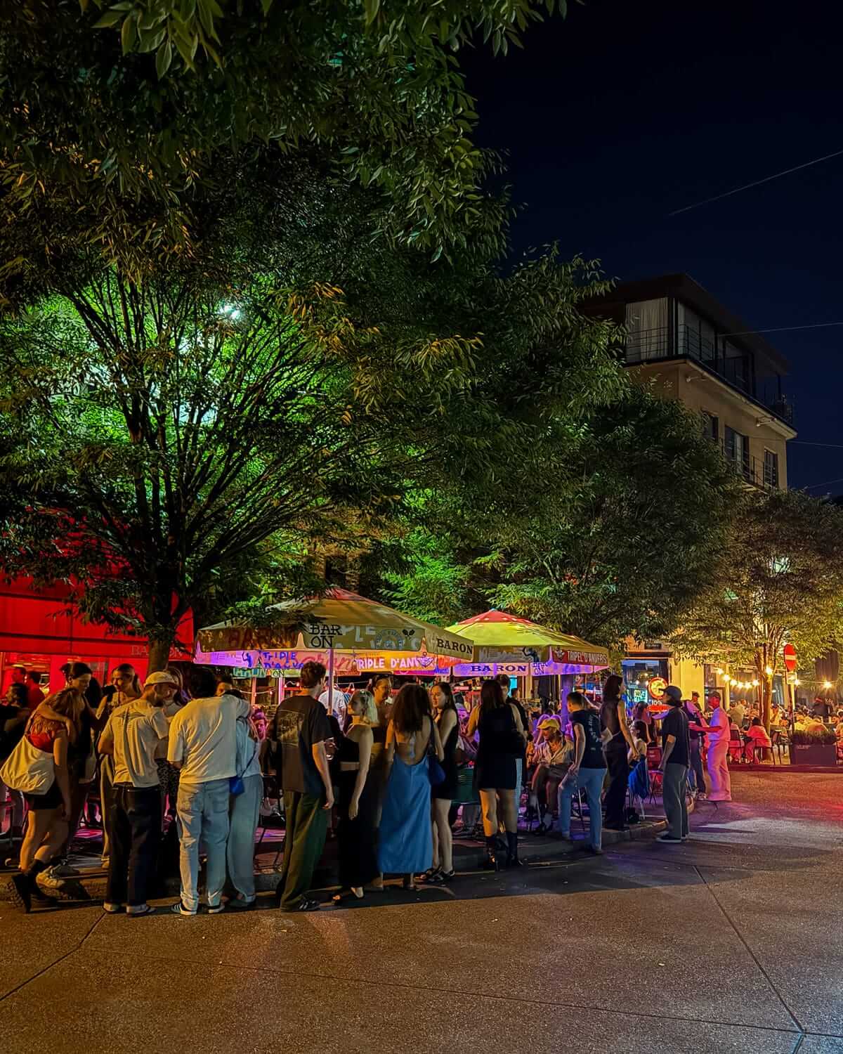 A lively young crowd gather outside at bars on Marnixplaats plaza, lit by colourful lights and surrounded by leafy trees at night.