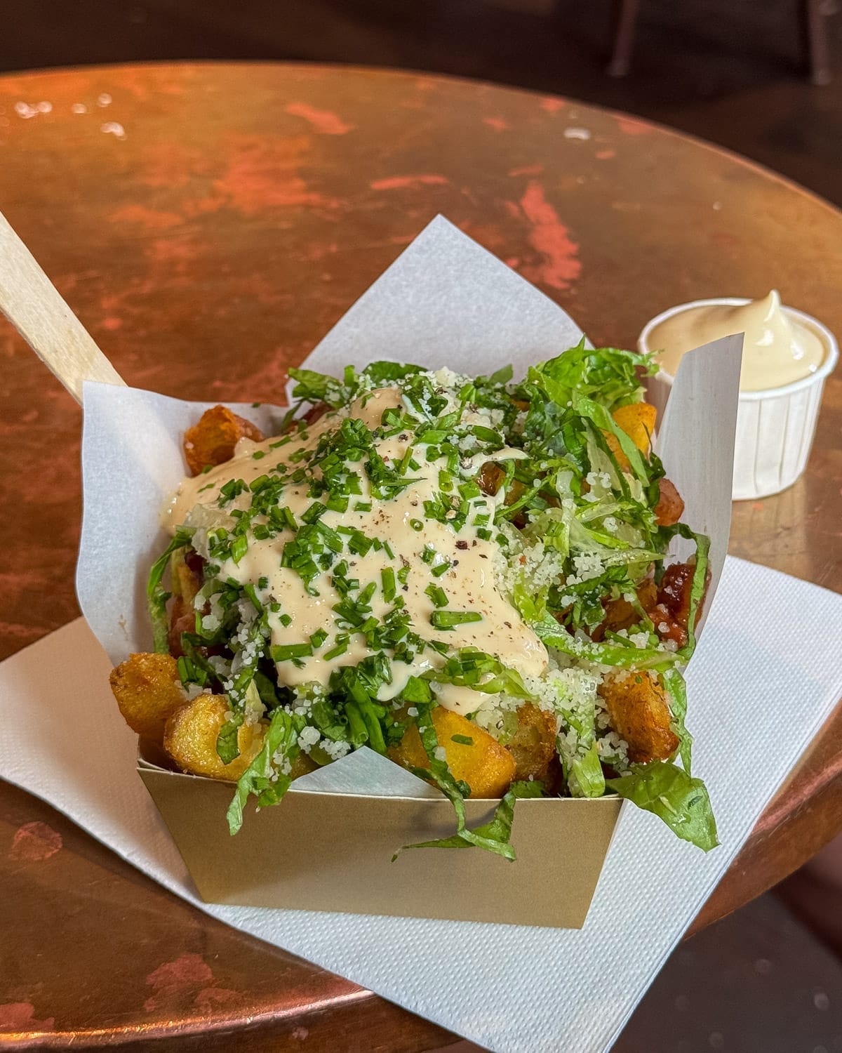 A metallic cardboard tray of crispy loaded fries from Frites Atelier in Antwerp topped with greens, chives and a creamy caesar sauce, served with mayo on the side.