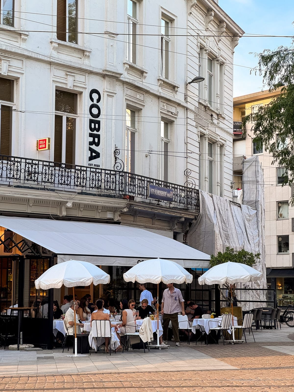 Stylish diners enjoy an elegant outdoor meal under white parasols on the corner terrace of Cobra, a stylish Antwerp restaurant.