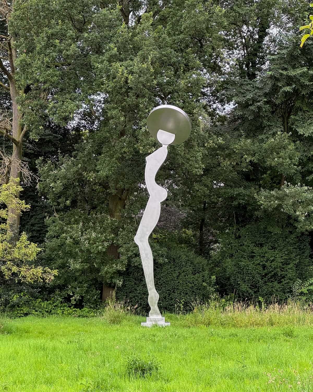 A curvy abstract sculpture of a woman with a wide hat stands in a grassy field against a backdrop of dense trees at Middelheim Museum in Antwerp..